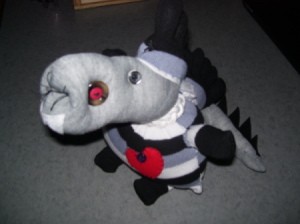 Cute long snouted creature made from a gray, black, and white striped sock. It has a spiky tail and a red bottle cap hanging around its neck.