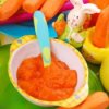 Dish of homemade carrot baby food. It is surrounded by fresh carrots and a ceramic bunny.