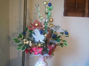 White vase with arrangement made up of silk poinsettas; silk vines; blue, gold, and silver christmas balls on sticks; and silver leaf decoration.