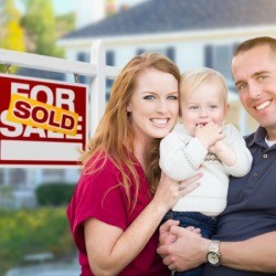 A happy couple with a baby outside a home with a for sale sign, marked sold.