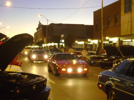 A classic car show with headlights on, at dusk.