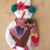 snowman door wreath wearing sweater, knit cap, gloves, and scarf
