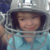 A smiling young girl with a very large Dallas Cowboys football helmet on.