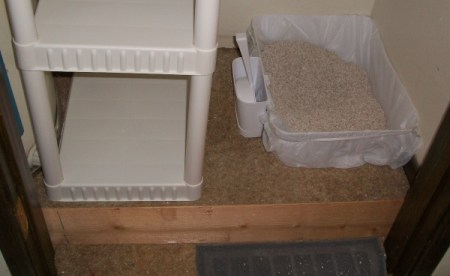 A shelving unit on the floor next to a water heater, with a cat litter pan next to it.
