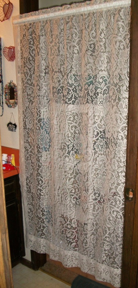 A lace curtain hiding the water heater and shelving unit.