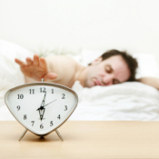 Man in bed hitting snooze button on an alarm clock.