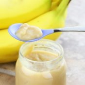 Jar of baby food with a spoon and bananas in the background.
