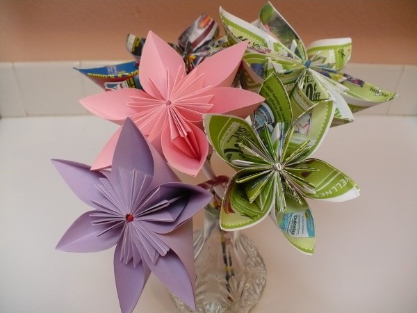 bouqet of paper flowers that includes a pink flower, a purple flower and 3 multi-colored flowers made from advertisements in a crystal vase