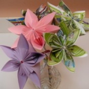 bouqet of paper flowers that includes a pink flower, a purple flower and 3 multi-colored flowers made from advertisements in a crystal vase