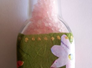 close up of bath salt bottle showing salts and oive paper wrapped around the bottle, white flower on right side gold dots along top edge of paper