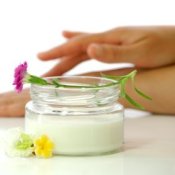 Jar of hand cream with flowers and woman's hands applying cream in the background.