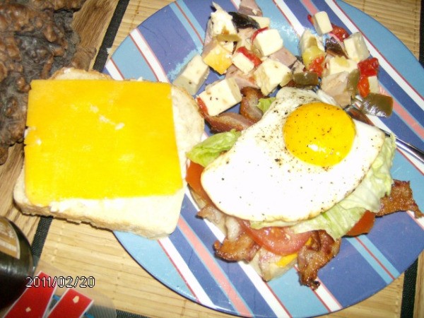 Stripped plate with two slices of bread. Slice on the right has bacon, lettuce, tomato, and fried egg on top; slice on left has cheese. Next to the bread is an antipasto salad.