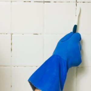 Blue gloved hand cleaning mold on shower walls with a toothbrush.