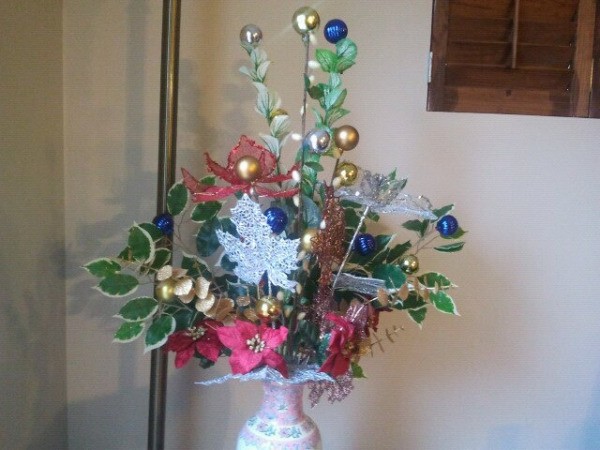 White vase with arrangement made up of silk poinsettas; silk vines; blue, gold, and silver christmas balls on sticks; and silver leaf decoration.