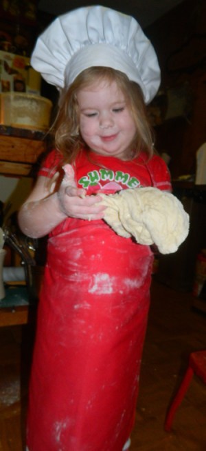 A little girl with a red apron and chef's hat kneading bread dough.