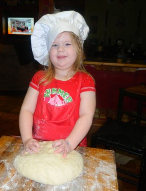 A little girl with a red apron and chef's hat kneading bread dough.