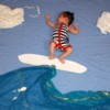 Baby lying on a sheet with other sheets and paper cutouts arrange to make it look as though he is surfing.