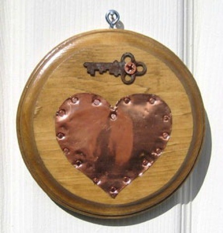 Copper heart on wooden circular plaque with a key attached above the heart.
