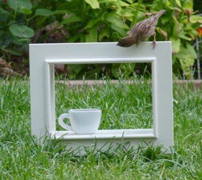 A sparrow on a white frame outside in the grass, with a teacup.