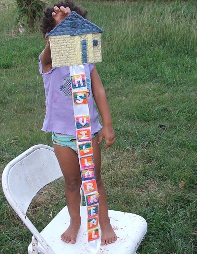 Little girl holding decoration consisting of house with blocks of letters hanging down.