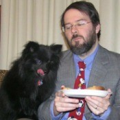 Black Pomeranian looking at man's food while licking her lips.
