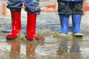 Two kids standing in the rain wearing colorful rain boots.