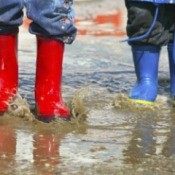 Two kids standing in the rain wearing colorful rain boots.