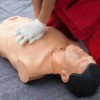Person in red scrubs kneeling and performing chest compressions on a plastic CPR "dummy"