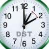 Close up of image of a white clock face with green border.  The clock has DST written on it and two hour hands exactly one hour apart with a red double ended arrow pointing at both hands indicating the hour changes forward and back by one hour