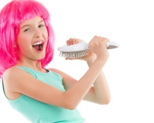 Young girl wearing hot pink wig singing into hairbrush against a white background