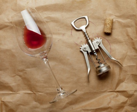 Image of wine glass on it's side with a small amount of wine, cork screw, and cork on a brown paper background with wine stains.