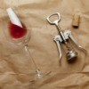 Image of wine glass on it's side with a small amount of wine, cork screw, and cork on a brown paper background with wine stains.