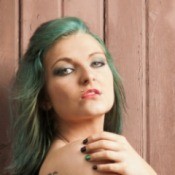 Woman with faded green blonde hair standing against a wood wall.  She is looking over her bare shoulder at the camera.