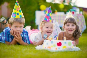 A little boy and two little girls wearing party hats laying their stomaches in the grass in front of a cake