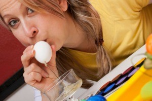 Blond woman blowing on one end of an egg.  The other end is over a bowl.  The egg contents are dripping into the bowl.