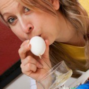 Blond woman blowing on one end of an egg.  The other end is over a bowl.  The egg contents are dripping into the bowl.
