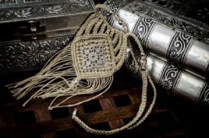 Intricate macrame (finger weaving) necklace resting against silver or pewter boxes on a dark wood surface.