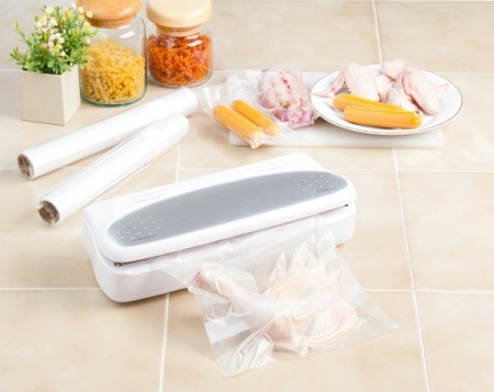 Vacuum sealer on tile counter with vacuum bag containing a chicken thigh.  Multiple other meat items such as hot dogs are off to the side and two rolls of unused vacuum bags are in the background.