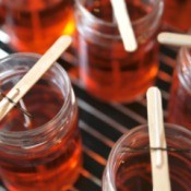 Several jars filled with hot red wax.  Sticks with wicks attached lay across the top of each jar with one end of the wick in the wax