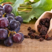 Close up of a bunch of red grapes next to a wooden scoop of raisins on a butcher block surface