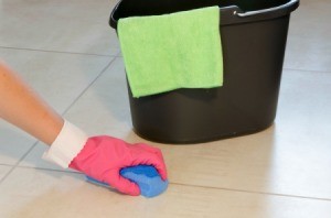 Hand wearing pink glove scrubbing white vinyl floor with blue sponge.  Black bucket with green cloth in the background