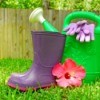 Hibiscus flower, goulashes, watering can, and gardening gloves displayed on grass with a wooden fence in the background.