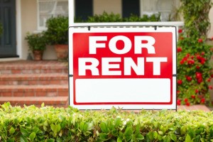 For Rent Sign hanging over a hedge with a home in the background