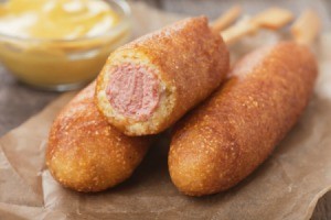3 Corn Dogs on a brown napkin with a bowl of mustard in the background.  One of the corndogs is half eaten.