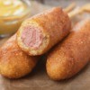 3 Corn Dogs on a brown napkin with a bowl of mustard in the background.  One of the corndogs is half eaten.