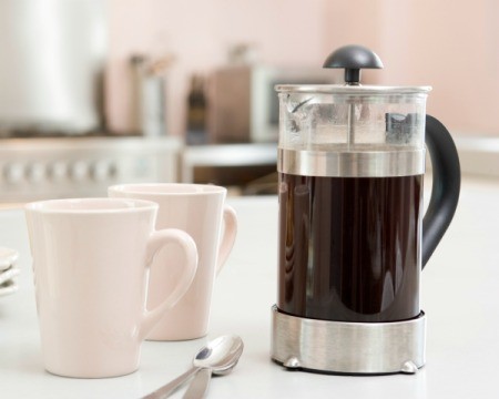 French press containing coffee, two white coffee cups, and two spoons on a white counter top