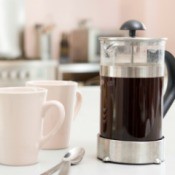 French press containing coffee, two white coffee cups, and two spoons on a white counter top