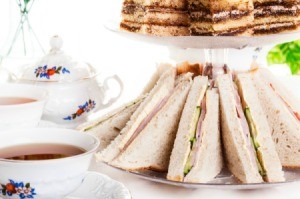 Plate of cucumber sandwich quarters arranged on tiered plate next to two china cups of tea and a teapot