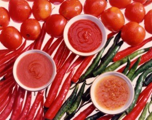 Bowls of tomato sauce, tomato sauce, and tomato puree' laying on top of fresh whole tomatoes and peppers on a white backgound