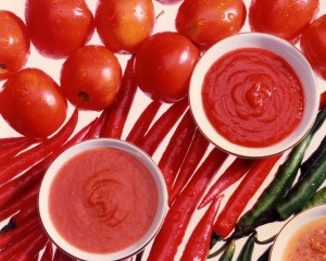 White bowls of tomato paste, tomato sauce, and salsa setting on a background of tomatoes and red and green peppers